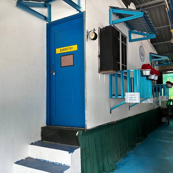 Pandangan sisi unit Dormstay dengan pintu masuk biru dan deretan beranda yang bersih di Kuala Selangor Cabin Camp (KSCC).