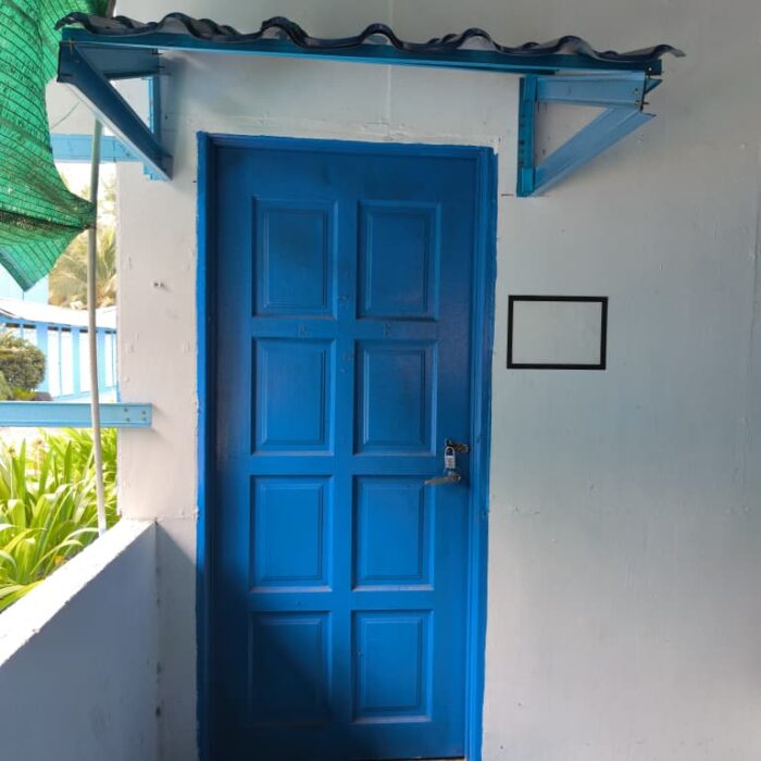 Bahagian luar Guest House dengan dinding putih, pintu biru, ampaian kain, dan tumbuhan hijau di Kuala Selangor Cabin Camp (KSCC).